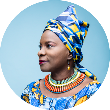 Side profile of Angélique Kidjo dressed in traditional patterned top and hat against a bright blue background.