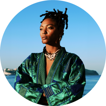 Bia Ferreira wears a green and blue satin patterned top and stares off to the distance in front of a coastal area.