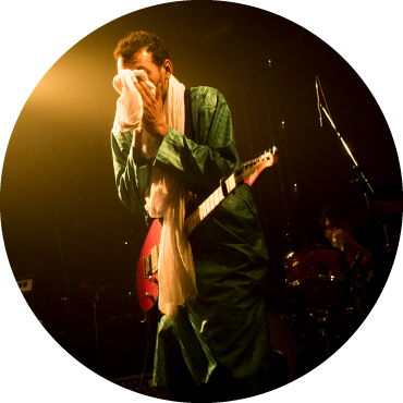Bombino wears green clothing and holds a towel to his face as he performs on stage with his guitar