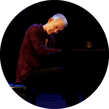 Brad Mehldau wears a red shirt and plays piano against a black backdrop
