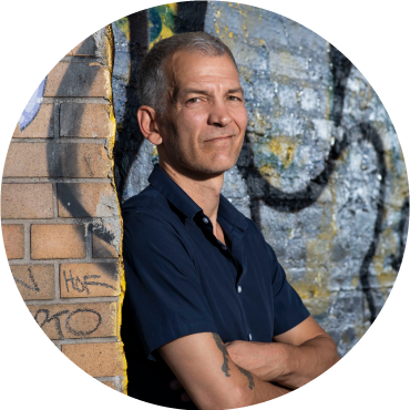 Brad Mehldau wears a blue shirt against a brick wall with his hands crossed
