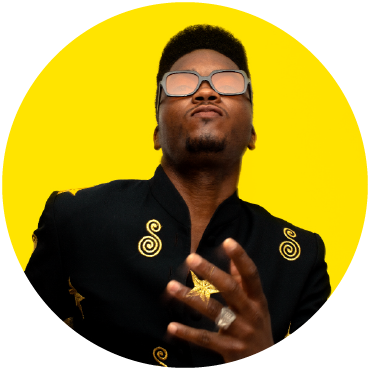 Performer Cimafunk from the chest up wearing an adorned black top and sunglasses, in front of a yellow background. Cimafunk holds a hand up to their chest..