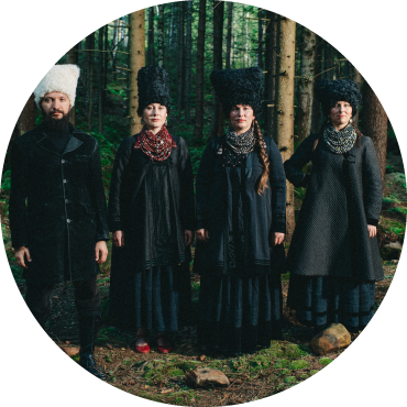 Four members of DakhaBrakha dressed in heavy fur hats and heavy black clothing stand in the woods together.