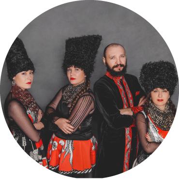 DakhaBrakha poses with their arms crossed wearing black and red traditional Ukrainian clothing in front of a gray background.