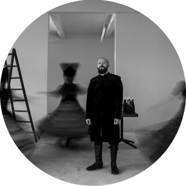 A black & white photo of a member of DakhaBrakha standing still while 3 women twirl around in dresses.