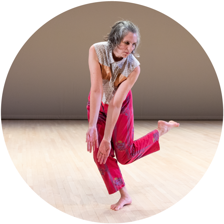 A dancer with chin length gray hair looks down to their left, bends over slightly with arms outstretched and left leg bent at the knee with foot in the air. The dancer wears red pants and a brown, blue, and white sleeveless button up shirt.