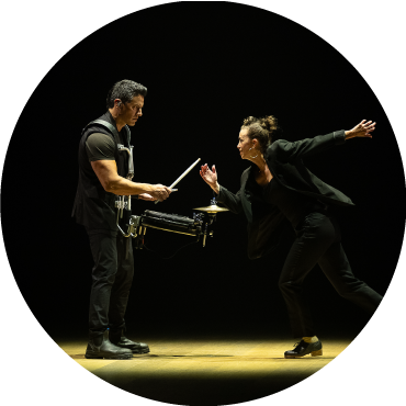 A female dancer busts a move and looks at a man playing an instrument on a dimly lit stage.