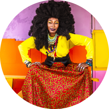 Fatoumata sits in front of a colored wall wearing a bright yellow long sleeve top, and an orange patterned skirt. Her hands are on her knees.