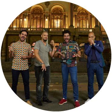 Harold López-Nussa's quartet poses on a street in front of Theatro Municipal in São Paulo, Brazil at night. The quartet is wearing jeans and pretending to play instruments. 