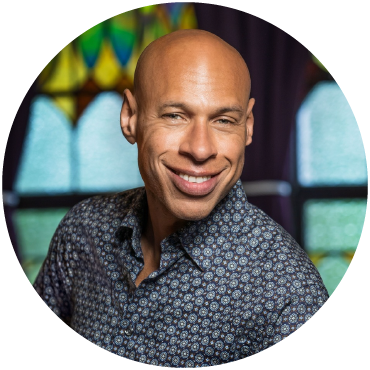 Joshua Redman dressed in a patterned blue button-down, smiles in front of a stained-glass background.