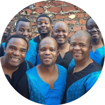 Seven members of Ladysmith Black Mambazo pose together smiling in front a brick background.