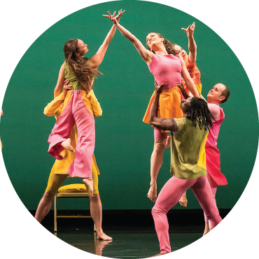 Two dancers dressed in pink and green cross hands on stage as they are held up by other dance members in front a green background.