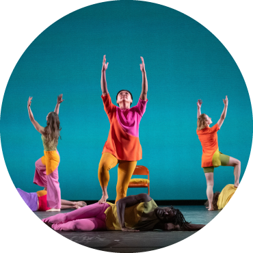 Three dancers stand with their arms raised over the bodies of other dancers on a stage against a turquoise background.