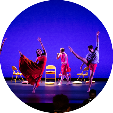 Two Dancers lift their legs on stage. In the background is another dance and three chairs.