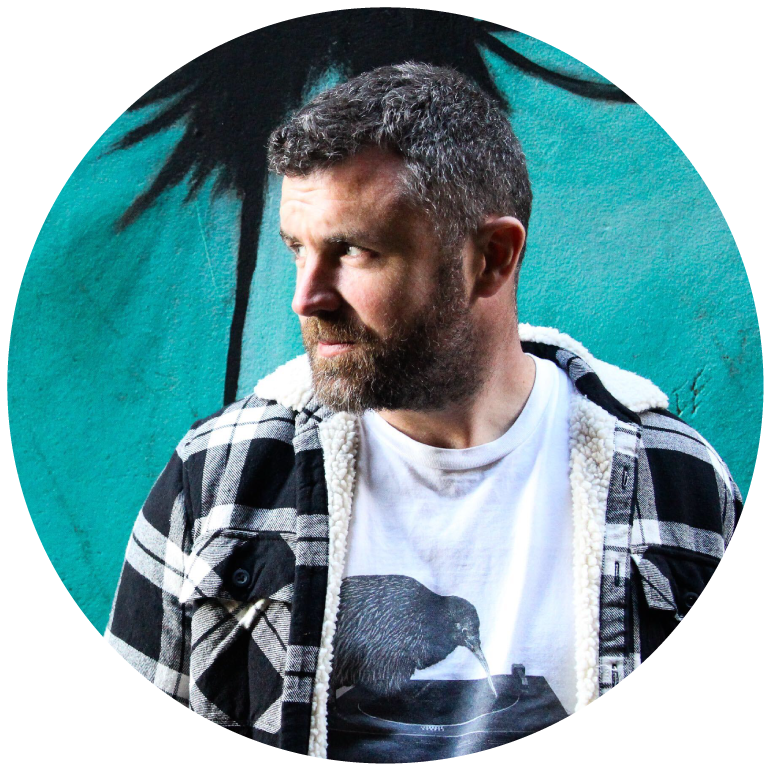 Mick Flannery wears a black and white flannel and looks off into the distance as he stands against a bright blue backdrop