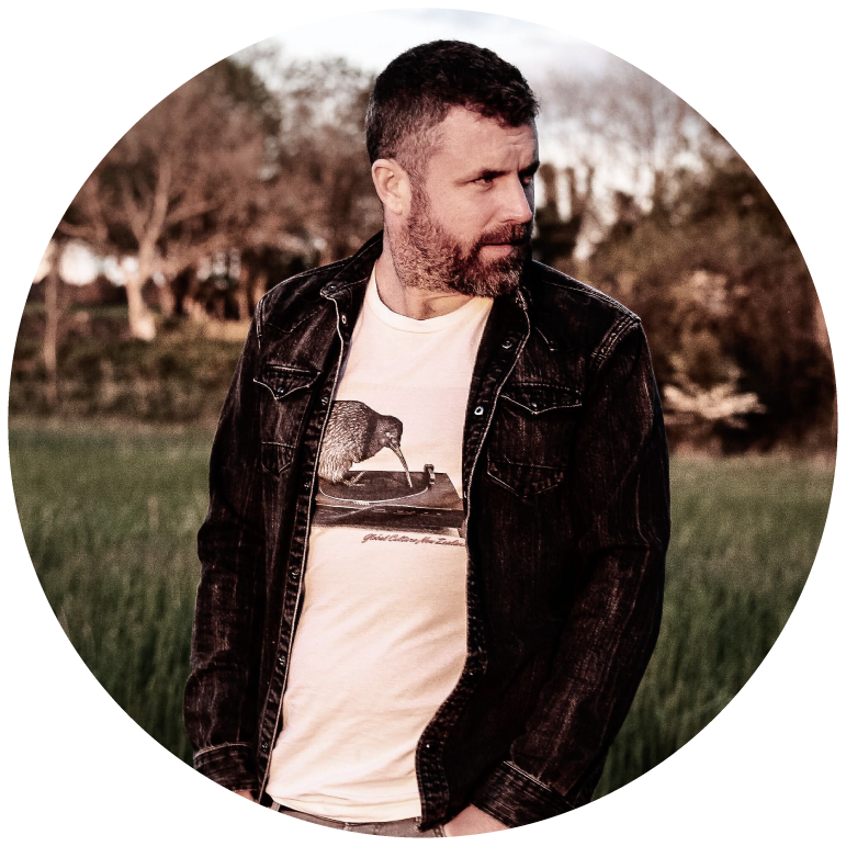 Mick Flannery wears a white shirt with a black denim jacket and looks to the side as he stands outdoors