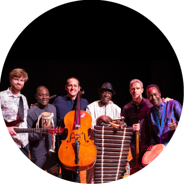 Mike Block holds a cello and Balla Kouyaté stands with his balafon as they pose with a larger group against a black backdrop