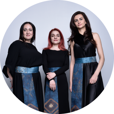 3 female members of the Naghash ensemble stand together wearing all black and turquoise patterned cloth worn on their waists.