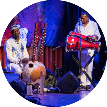 Seckou Keita dressed in white plays the kora on stage next to a percussionist playing a large red drum.