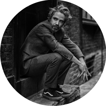 Patrick Watson sits against a brick wall dressed in a suit and looks forward with his hands on his knees
