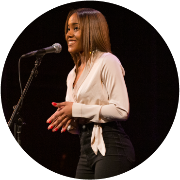 A circular image of a person standing in front of a microphone. They have medium length, straight, light brown hair and are wearing a white blouse and black pants.