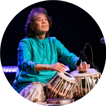 Zakir Hussain wears turquoise and looks off as he plays the tabla drums.