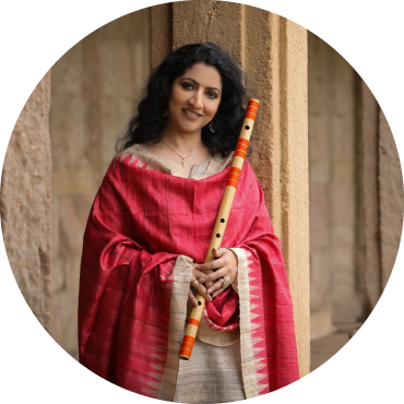 A smiling woman wears red and beige traditional Indian clothing and holds the the bansuri (flute)