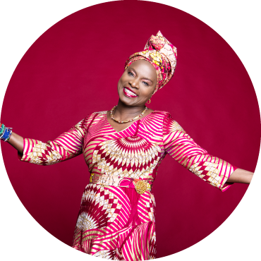 Angelique stands against a pink background wearing a beautiful dress and headscarf with a big smile and her arms outstretched
