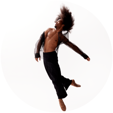 A dancer with long curly hair dressed in black leaps up into the air against a white background. 