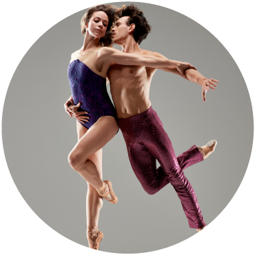 In a gray background two dancers leap as they hold each other. One dancer wears blue and the other dance wears purple pants.