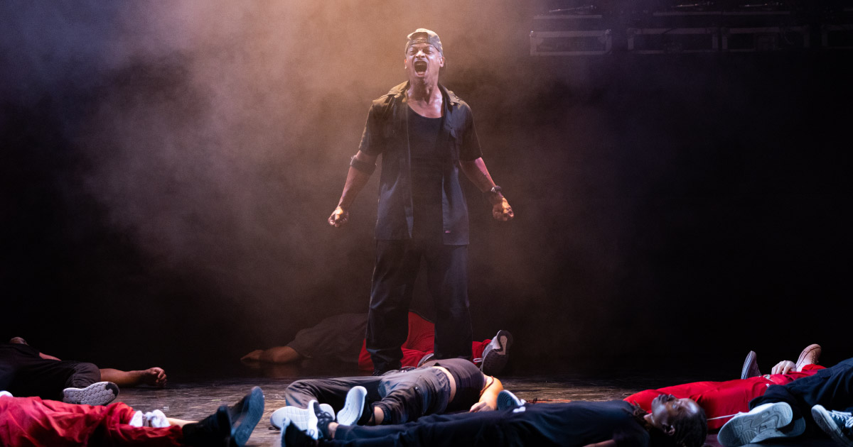 A dancer stands, fists clenched and screaming, surrounded by other dancers laying on the floor