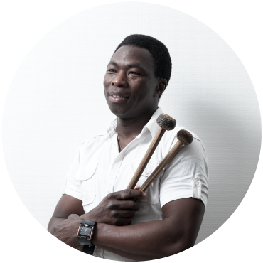Aly Keïta wears a white shirt and holds the mallets of a balafon against a white background.