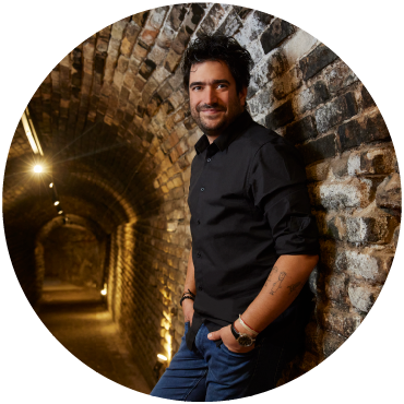 Harold López-Nussa leans against the stone interior of a dimly-lit tunnel wearing a black button-down and blue jeans.