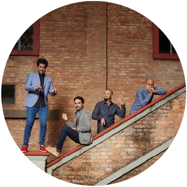 Harold López-Nussa's quartet pretends to play instruments by the stairs of a brick building. Two men are on top of the staircase posing, two men are behind the staircase wearing blue shirts. 