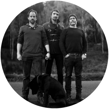 The Langan Band trio stand together looking into the camera in a wooded area, a black dog sits in front of them. The photo is black and white.