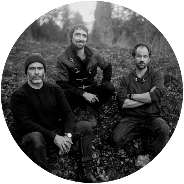 The Langan Band trio sit down together in a wooded area, looking into the camera. The photo is black and white.