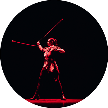 A performer of Malevo swings two long objects on a stage lit by red lighting.