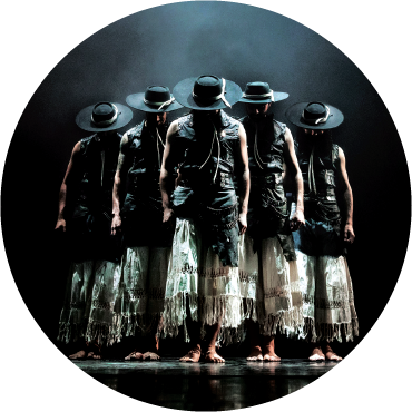 Five performers of Malevo stand on stage barefoot together with their heads down and wearing black tops and white skirts against a black background.