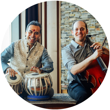 Sandeep Das smiles and plays the tabla while seated next to Mike Block holding a cello.