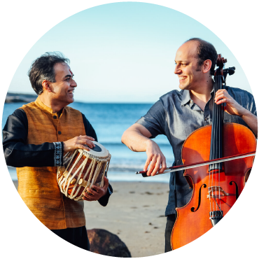 Sandeep Das playing the tabla and Mike Block playing the cello stare into each other eyes at the beach.