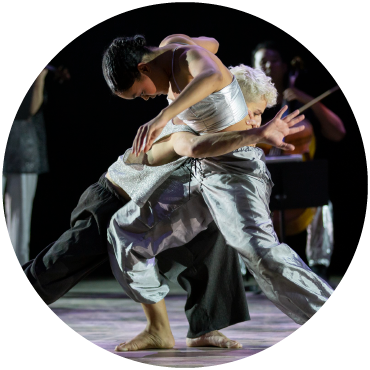 Two dancer extend their legs into each other on stage as a musician plays the cello in the background.