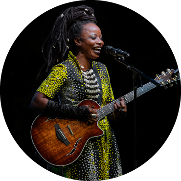 Singer Natu Camara wears a yellow, black, and white-dotted dress as she a brown guitar and sings into a microphone.