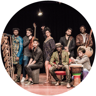 Sidy Samb poses with an assemble of performers holding instruments on a wooden floor. A flamenco dancer stands in the middle wearing black holding an arm up.