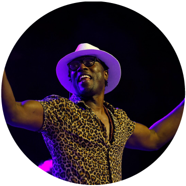 Sidy Samb exclaims on stage wearing cheetah print and a white fedora. Sidy samb is lit by purple lighting.