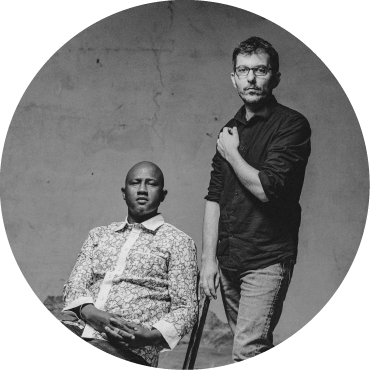 Black & white photo of Abalye Cissoko sitting next to Cyrille Brotto who stands.