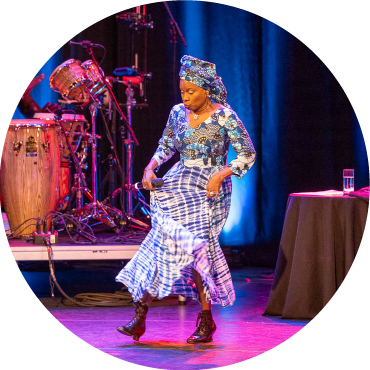 Angélique Kidjo lifts her vibrantly patterned dress as she dances on a stage.