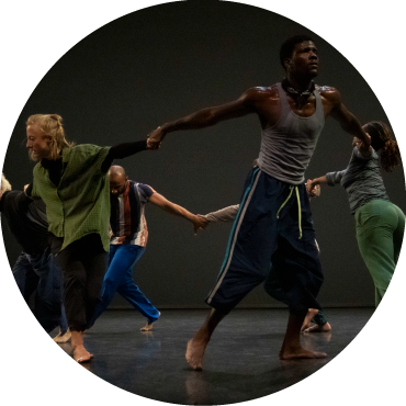 Five members of David Dorfman Dance dressed in black, green, and grey hold their hands together in a circle with their backs facing each other.