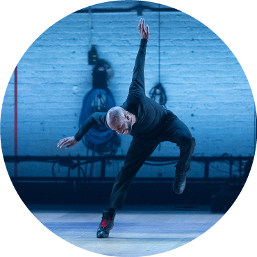 A male dancer stomps his left foot down and throws his arm in the air while on a stage lit by subtle blue lighting.