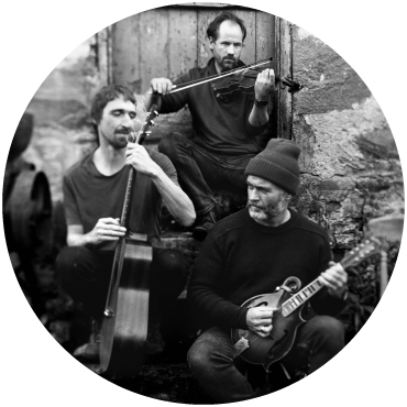 The Langan Band trio sit in front of the steps of an old building playing a fiddle, a bass guitar, and strumming a guitar. The photo is black and white. 
