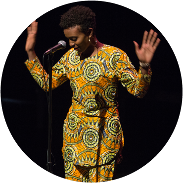 A circular image of a person with short black curly hair wearing a pattered set of a yellow shirt and matching pants. They are smiling while looking at the ground and holding their hands in the air. 